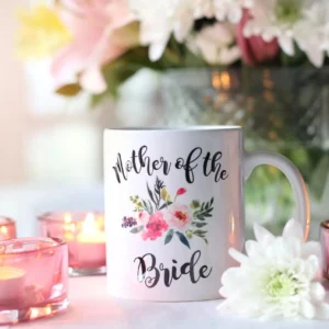 ספל קרמיקה לבן עם הכיתוב Mother of the Bride ואיור פרחים, מונח על שולחן לצד נרות דולקים בפמוטי זכוכית וזרי פרחים